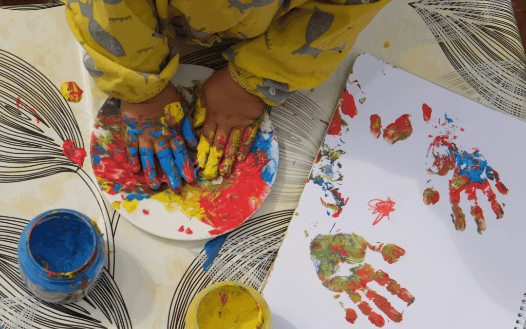 Child's hands covered in bright primary paint resting on a plate while a paint-splotched notebook lies nearby during a messy art activity.
