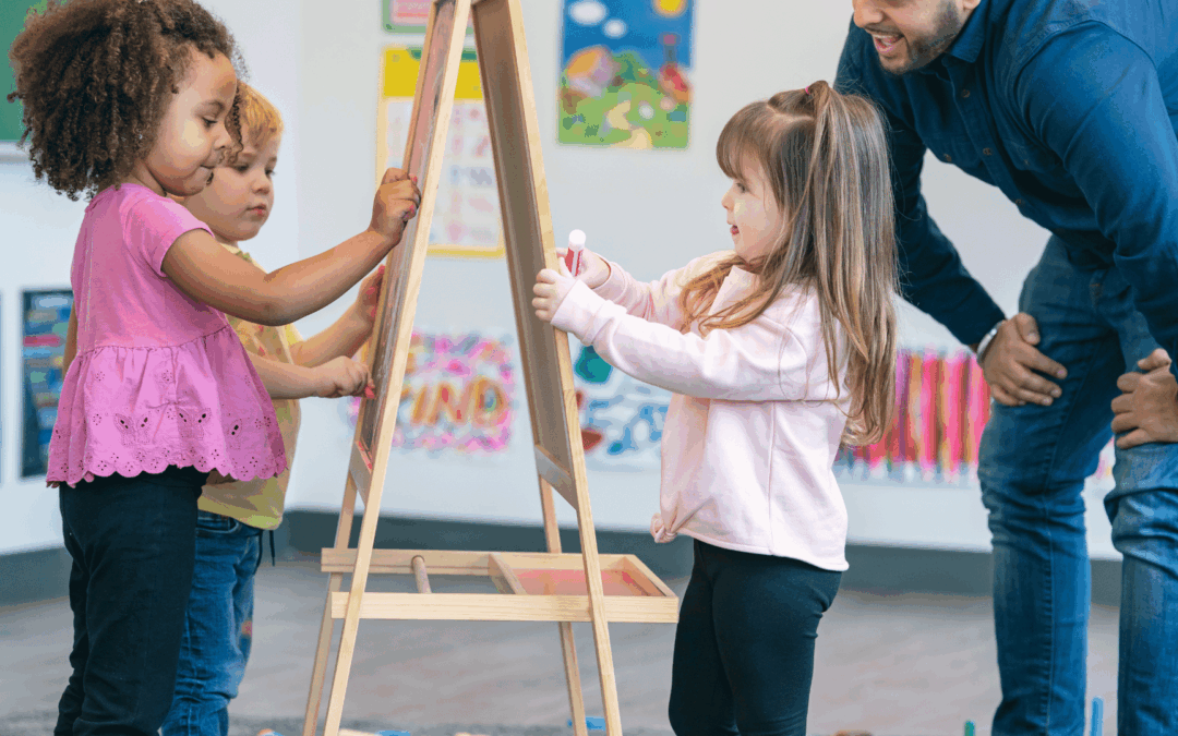 Children draw on easels at a classroom activity, guided by an adult supervisor nearby.